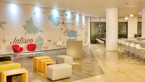 Un moderno vestíbulo de hotel con un mapa de Jalisco, México, zonas de estar y un bar/lounge con sillas blancas.