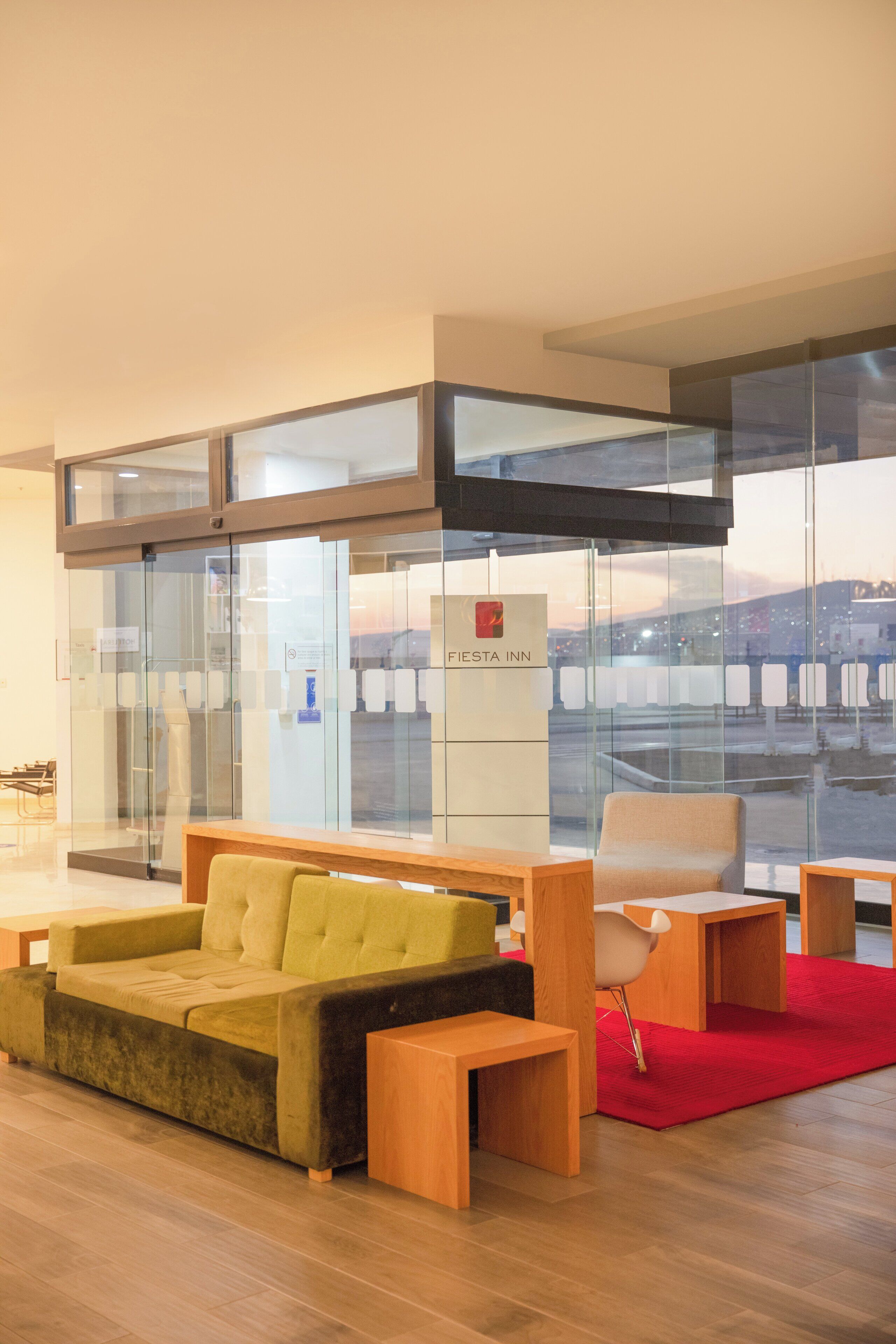 A modern hotel lobby with a sofa, chairs, a red rug, and a glass wall with a city view.