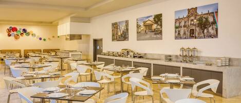 A spacious dining area with multiple tables and white chairs, a long counter with kitchen appliances, and wall-mounted decorative plates.