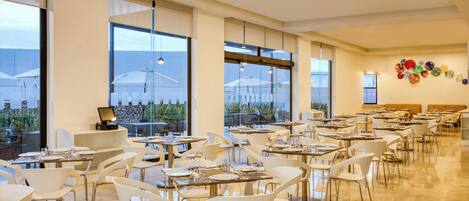 Un comedor amplio con varias mesas y sillas, ventanas grandes que ofrecen vistas al exterior, y una pared decorada con platos coloridos.