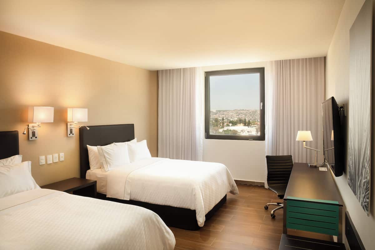A hotel room with two beds, a desk, a chair, and a large window offering a city view.