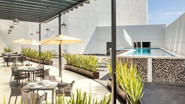 Un patio al aire libre con mesas, sillas, sombrillas y una piscina.