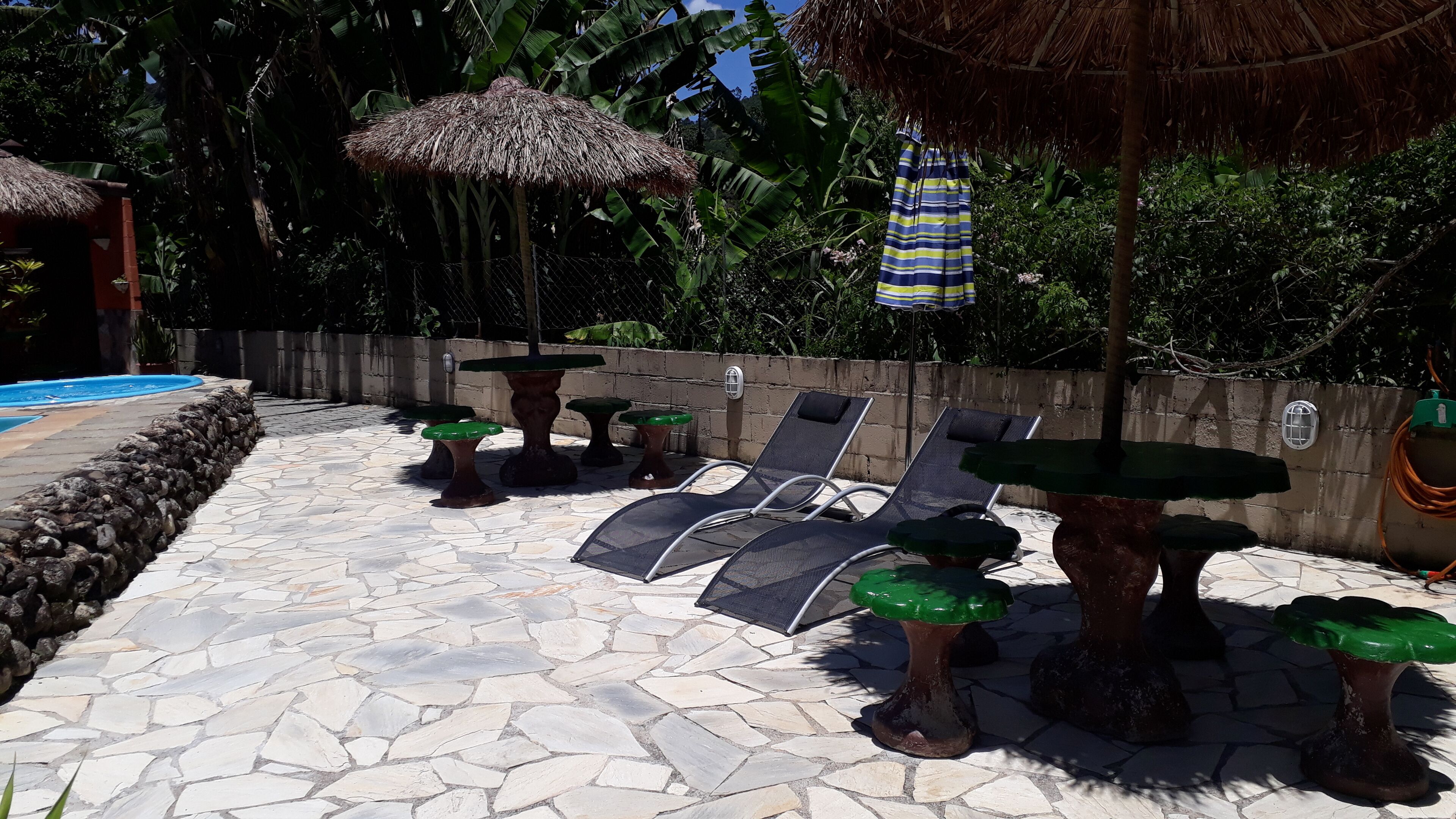 Un área junto a la piscina con suelo de piedra, sillones de descanso y sombrillas de paja.
