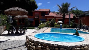 Ein Schwimmbecken mit Steinrand, umgeben von einer Terrasse mit Tischen und Sonnenschirmen.
