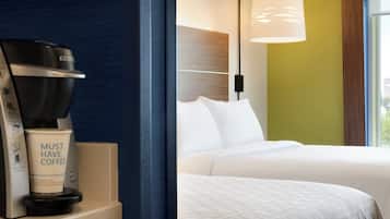 Una habitación de hotel con cafetera, una taza que dice 'NECESITO CAFÉ' y una cama con ropa blanca.