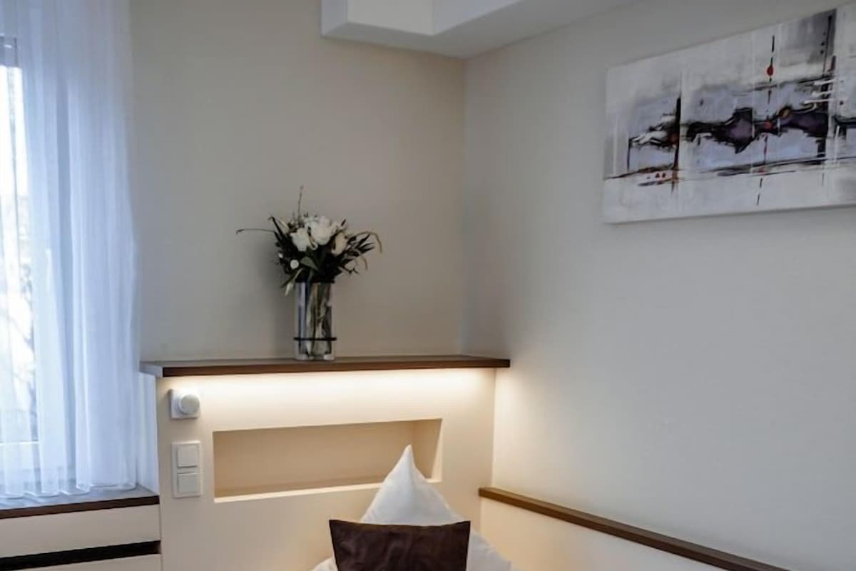 Un lit simple avec une tête de lit, une table de chevet avec une lampe, une œuvre d’art accrochée au mur et une fenêtre avec des rideaux.