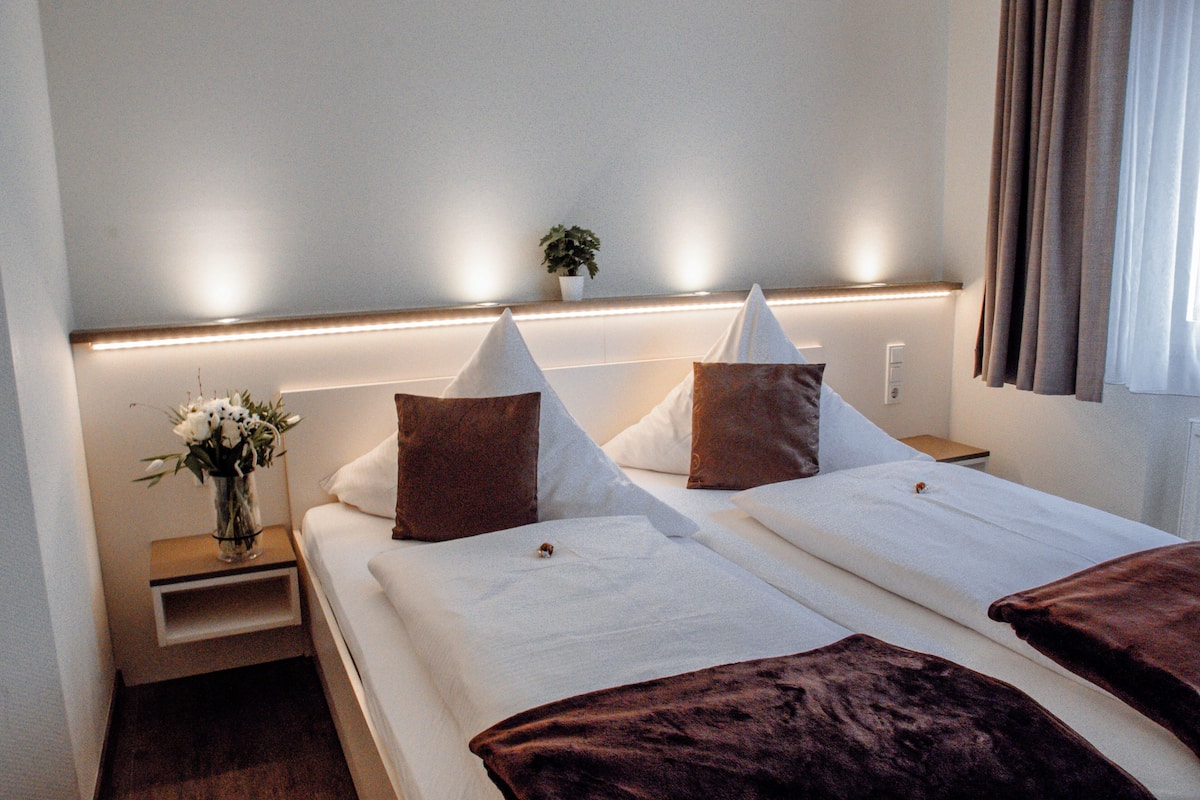 Ein Hotelzimmer mit zwei Betten, einem Nachttisch mit Blumenvase und einem Beistelltisch.