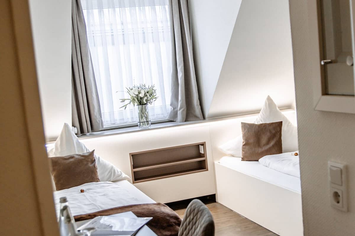 Ein Hotelzimmer mit Bett, Schreibtisch, Stuhl, einem Fenster mit Vorhängen und einem kleinen Tisch mit Blumen.