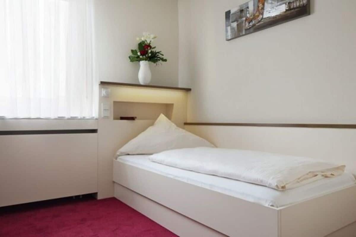 Ein kleines, ordentlich eingerichtetes Schlafzimmer mit einem Einzelbett, einem Nachttisch, einem an der Wand befestigten Regal mit einer Blumenvase und einem gerahmten Bild.