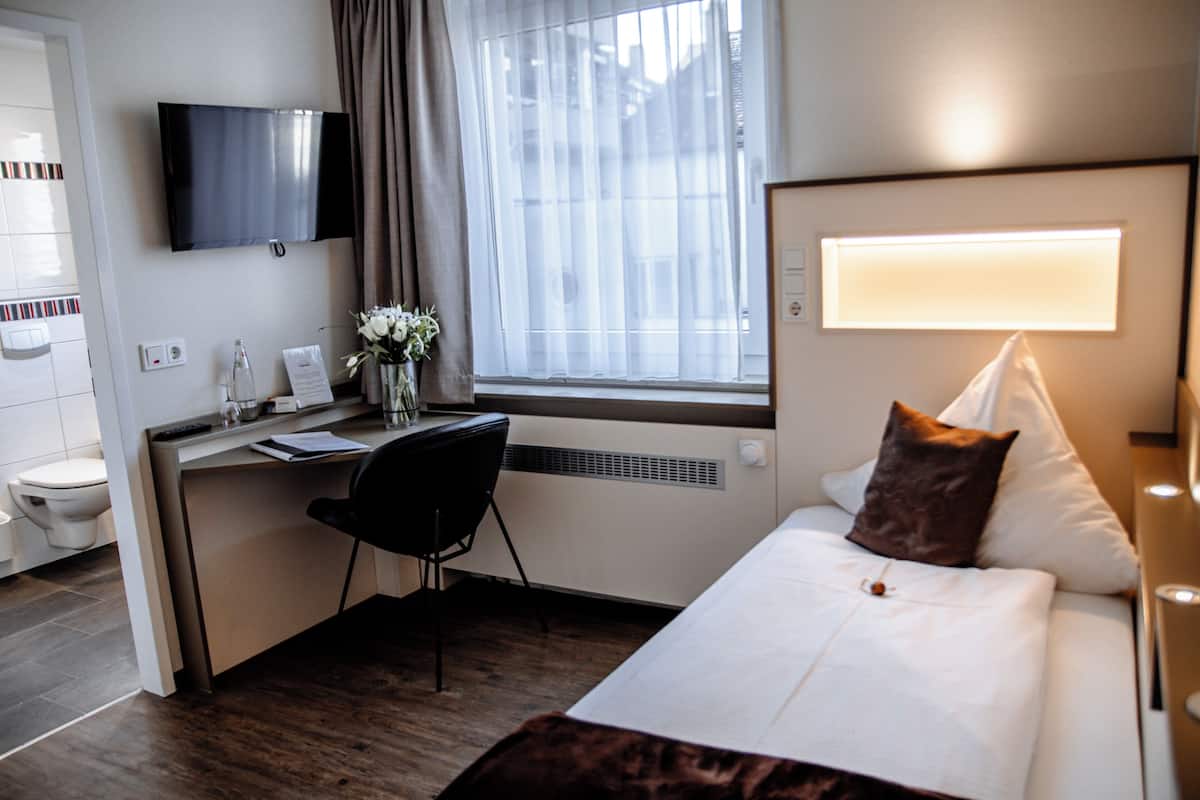 Ein Hotelzimmer mit einem Bett, einem Schreibtisch, einem Stuhl, einem Fernseher, einem Fenster mit Vorhängen und einem Badezimmer mit Toilette und Waschbecken.