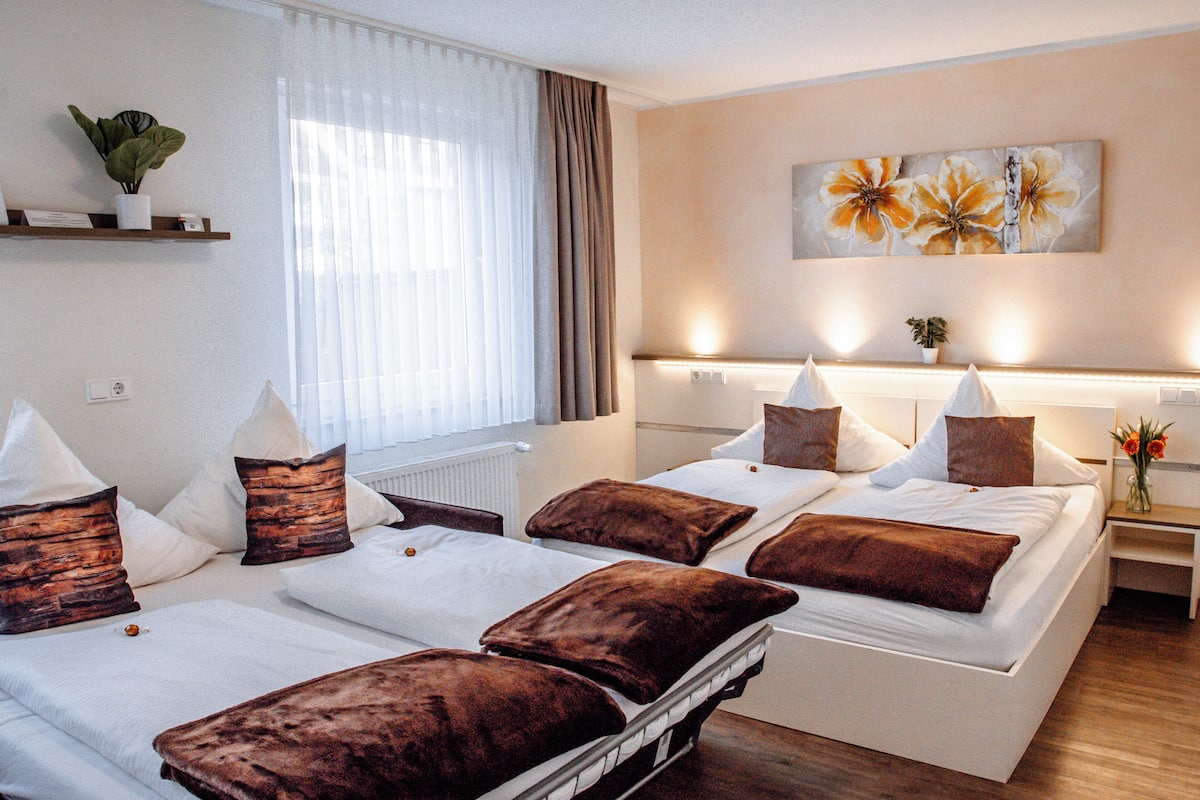 Ein Hotelzimmer mit zwei Betten, jeweils mit braun-weißer Bettwäsche, einem großen Fenster mit durchscheinenden Vorhängen, einem Wandbild und einer kleinen Pflanze auf einem Regal.