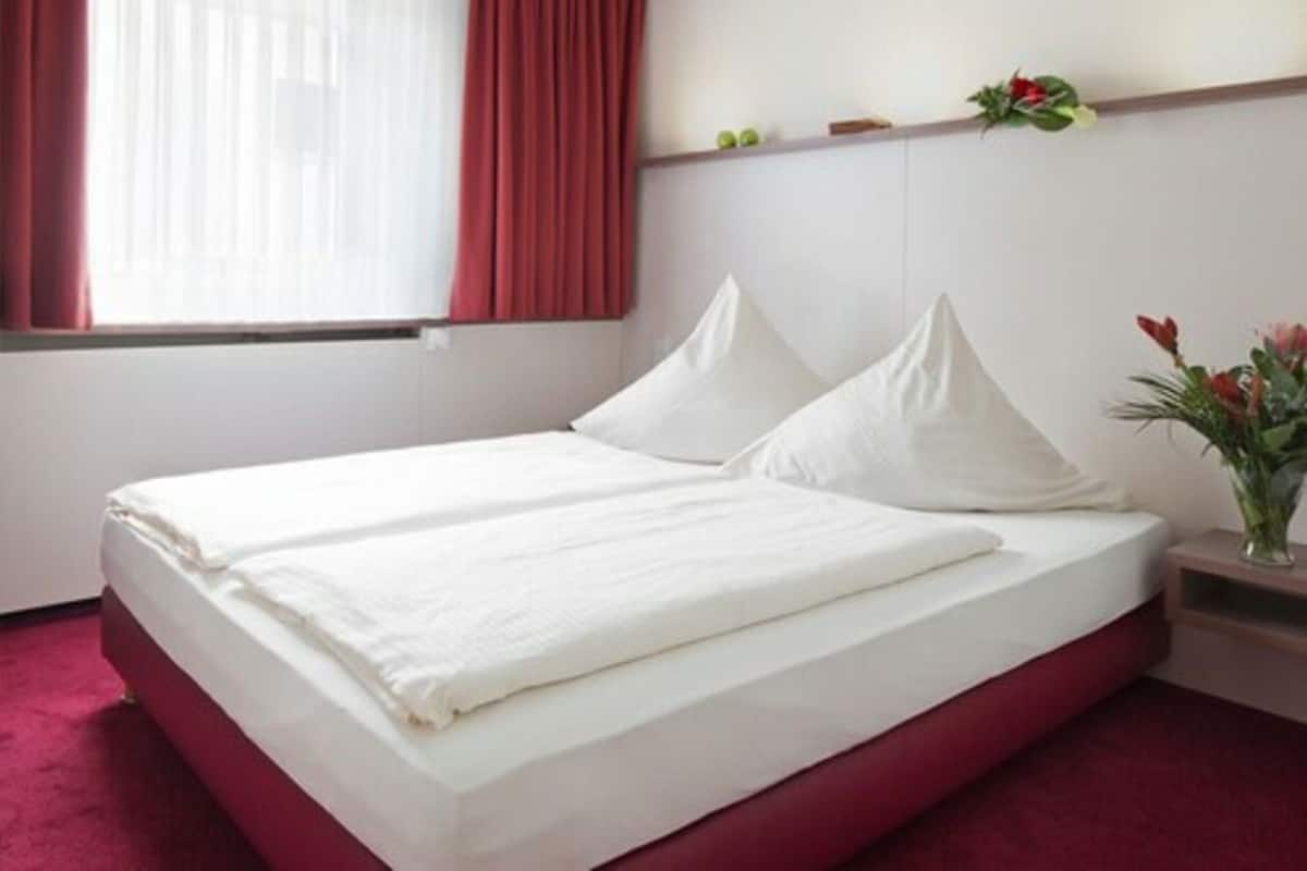Ein Bett mit weißer Bettwäsche, eine rote Akzentwand und ein kleiner Nachttisch mit einem Blumenstrauß.