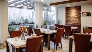 Een restaurant met meerdere tafels en stoelen, grote ramen met uitzicht op bomen, en een decoratieve stenen muur.