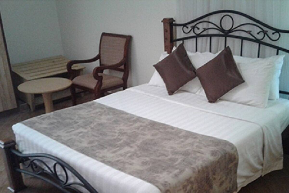 Een opgemaakt bed met hoofdbord, een stoel, een klein rond tafeltje en een nachtkastje met een lamp.