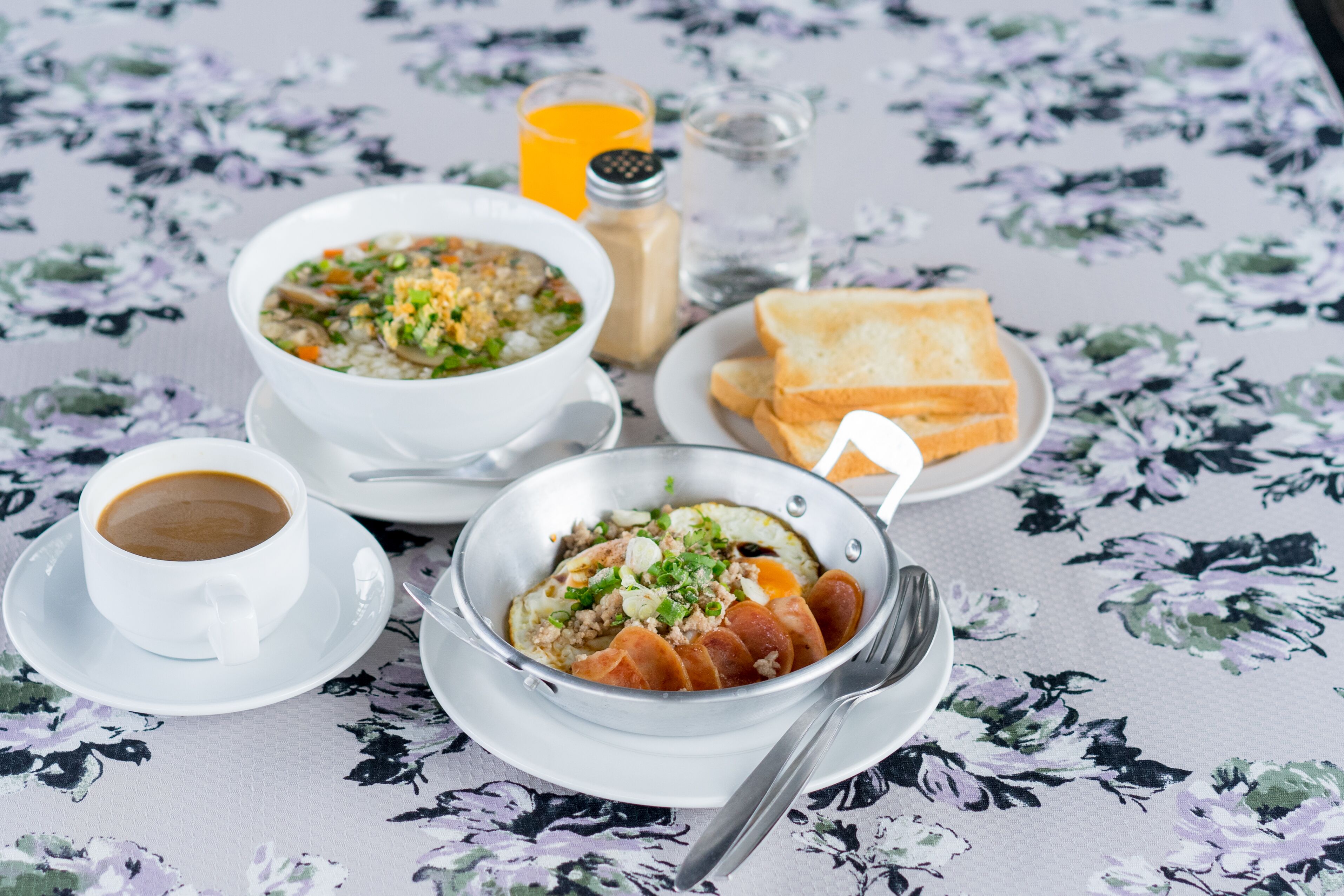 A table with a floral tablecloth, two bowls of soup, a plate of toast, a cup of coffee, and a glass of orange juice.