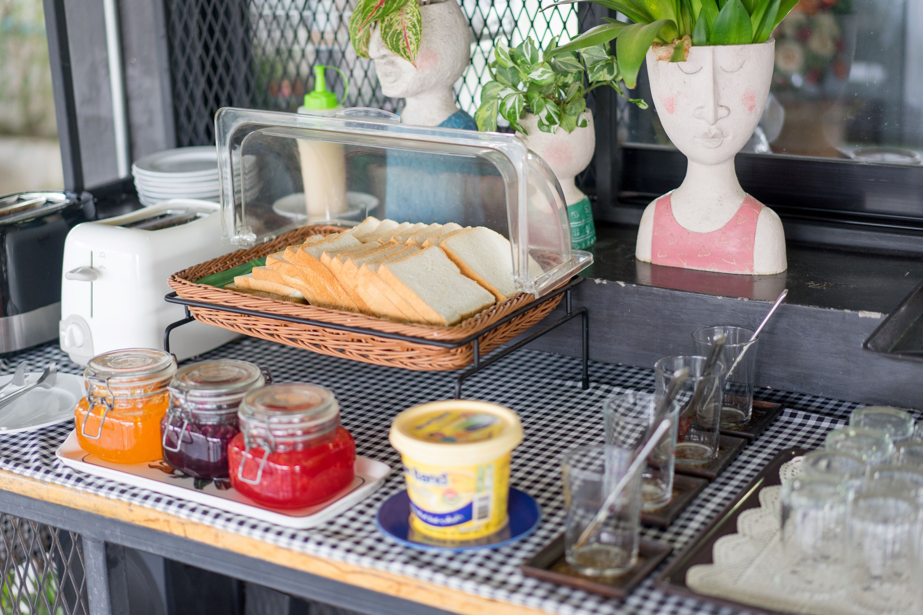 A breakfast setup with a basket of bread, jars of jam, and a yogurt container on a checkered table.