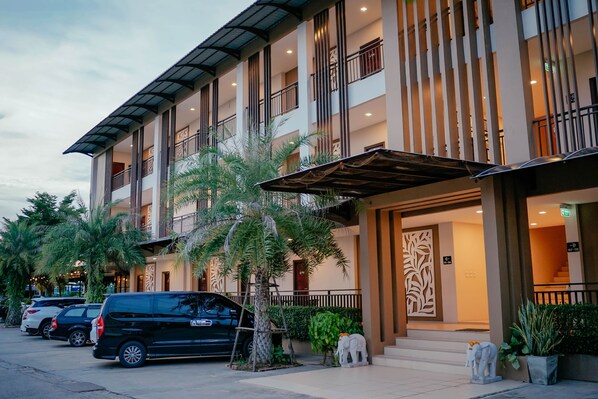 Un hotel de varios pisos con un diseño moderno, que cuenta con balcones, una entrada cubierta y un estacionamiento con varios automóviles.
