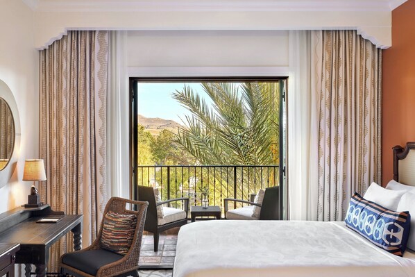 A hotel room with a large bed, a desk with a lamp, a chair, a small table with a vase, and a balcony with a view of palm trees and mountains.