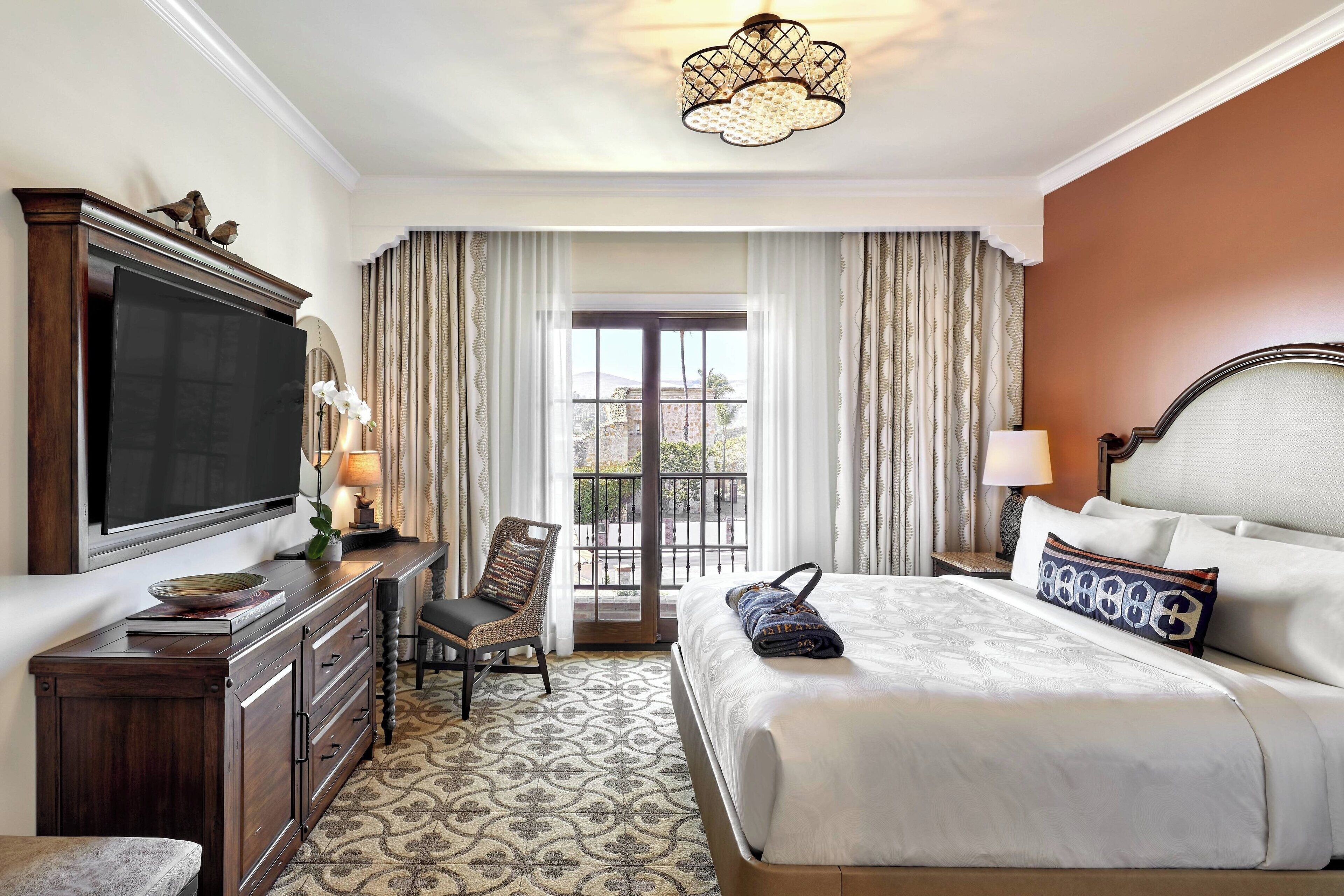 A hotel room with a large bed, a television on a wooden cabinet, a chair, a desk, and a view of the outdoors through a window.