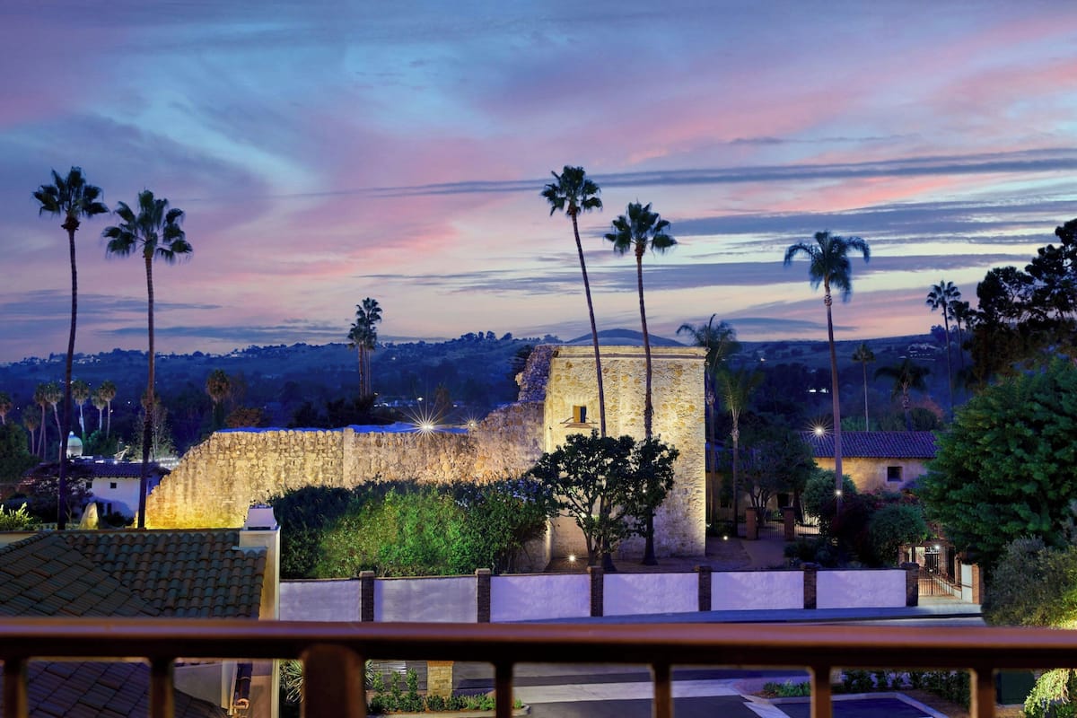 A historic stone building with a fortified wall, surrounded by palm trees and a sunset sky.