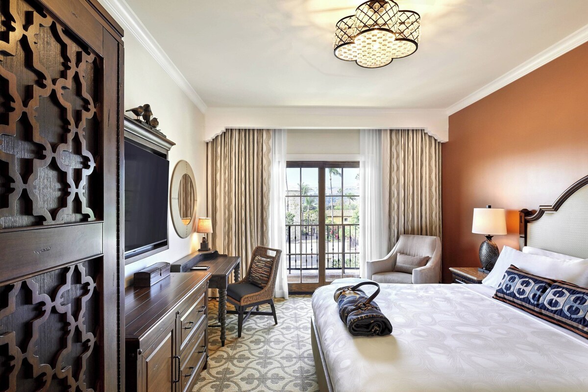 Habitación de hotel con una cama grande, un escritorio, una silla, un televisor y un balcón con cortinas.