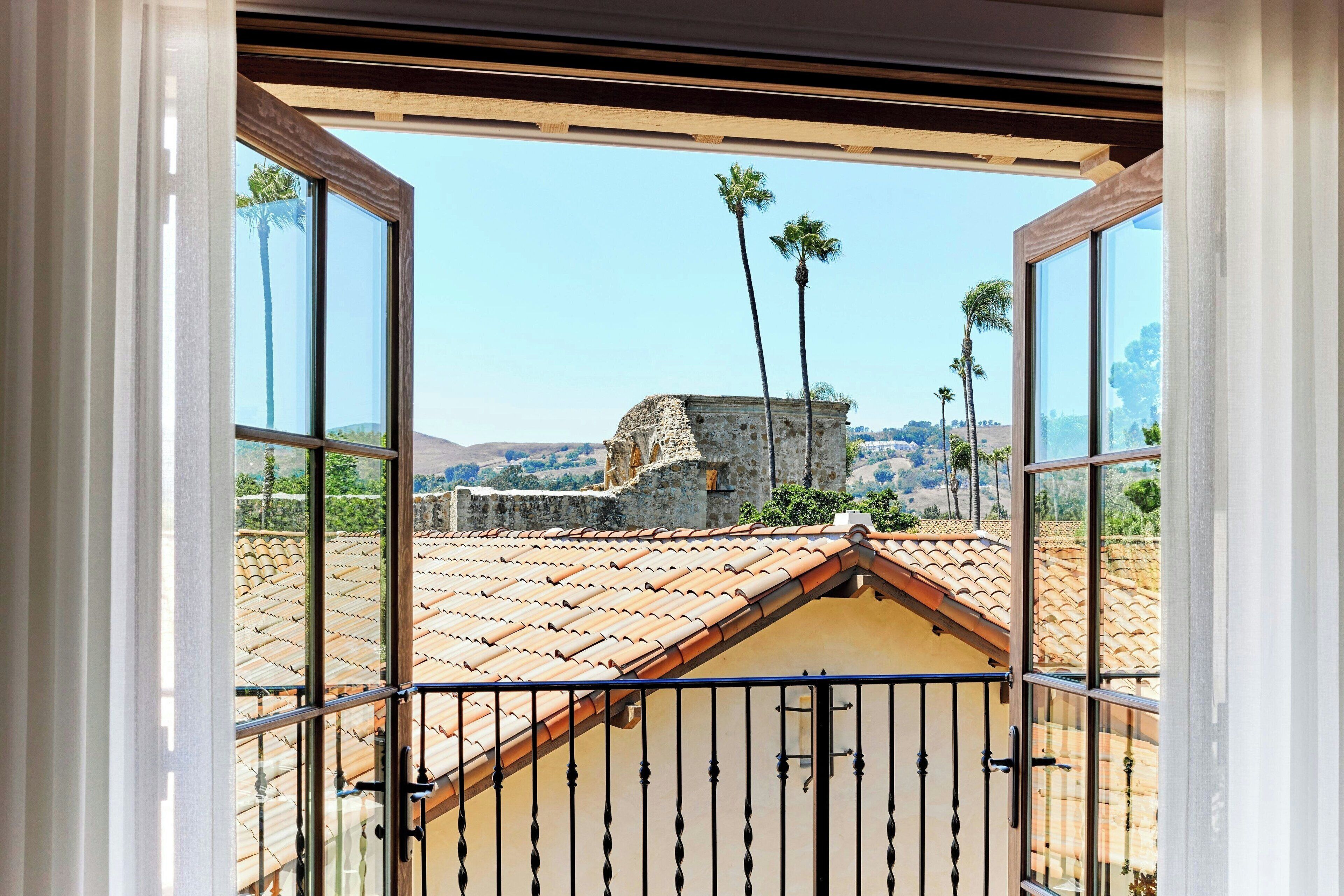 A balcony with a view of a tiled roof, palm trees, and a stone structure.