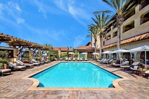 A swimming pool with lounge chairs and umbrellas, surrounded by palm trees and a building with a tiled roof.