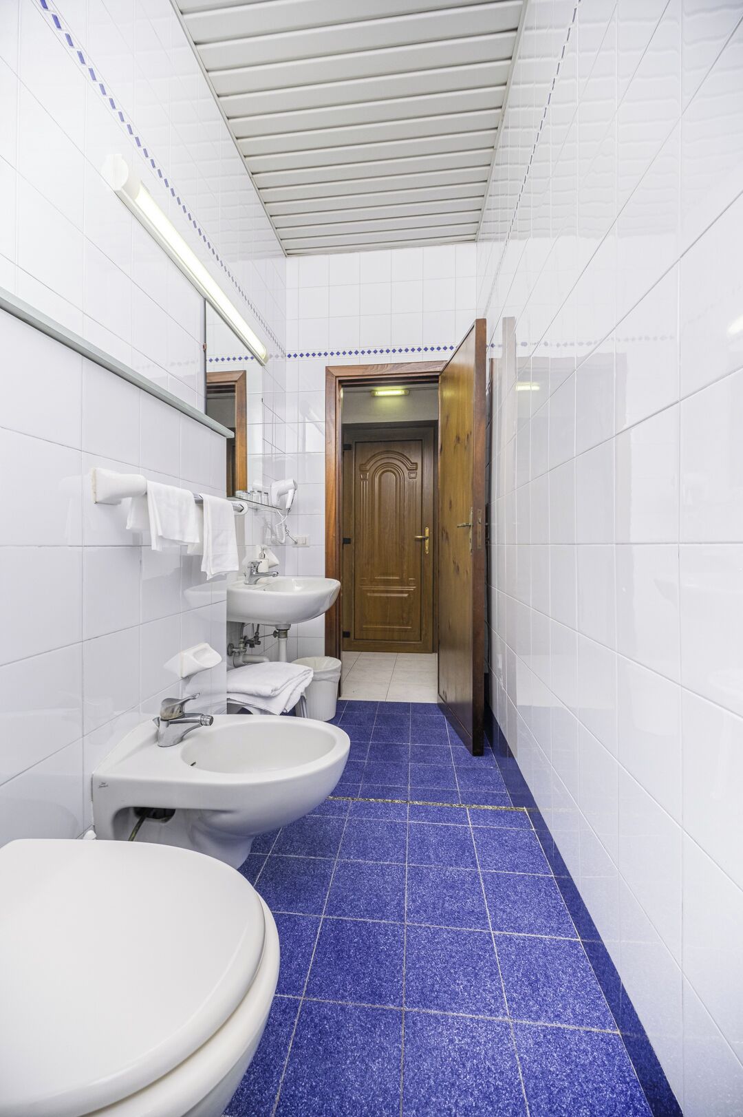 Une salle de bain avec des murs carrelés de carreaux blancs, un sol carrelé de carreaux bleus, une porte en bois et un miroir.