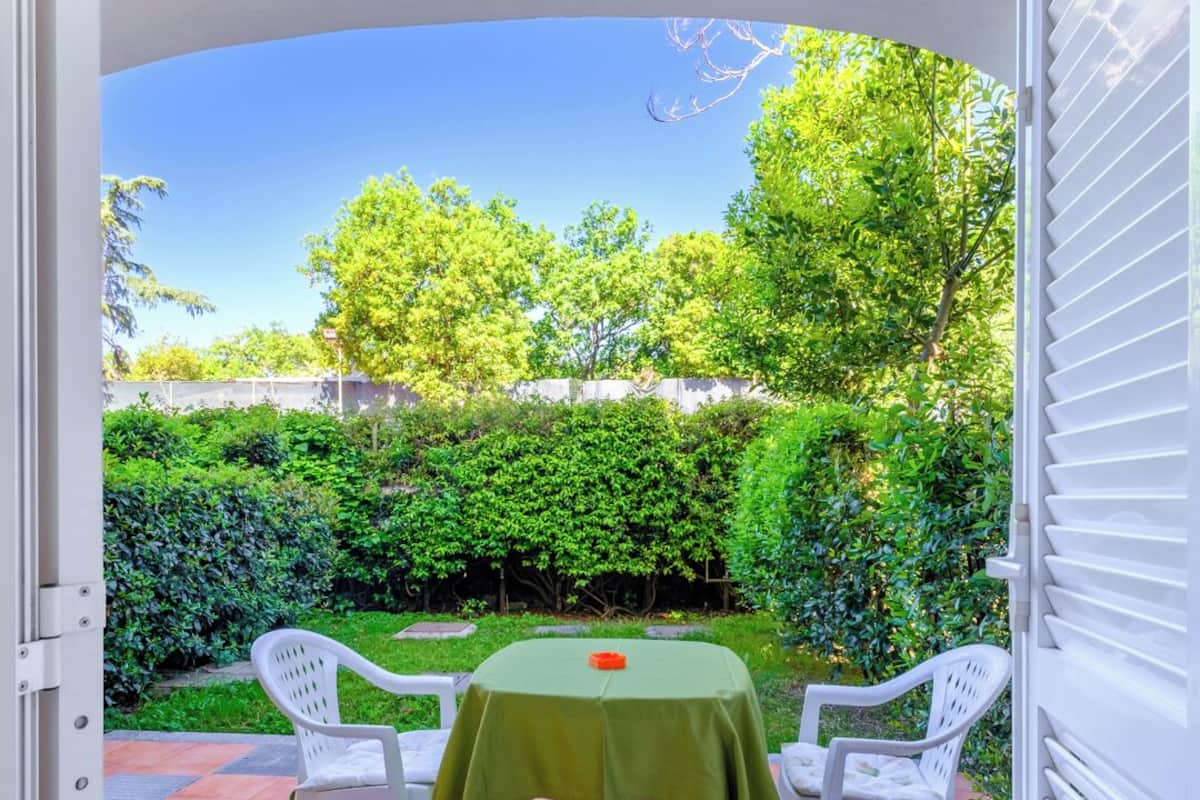 Eine offene Terrasse mit Tisch und zwei Stühlen, einer grünen Tischdecke und Blick auf einen Garten mit Bäumen und einer Hecke.