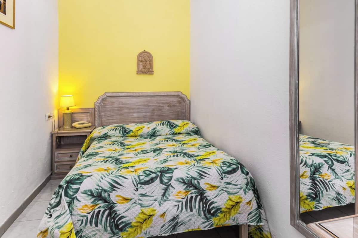 Ein kleines, helles Schlafzimmer mit einem Einzelbett, einem Nachttisch, einem Spiegel und einer gelben Wand.