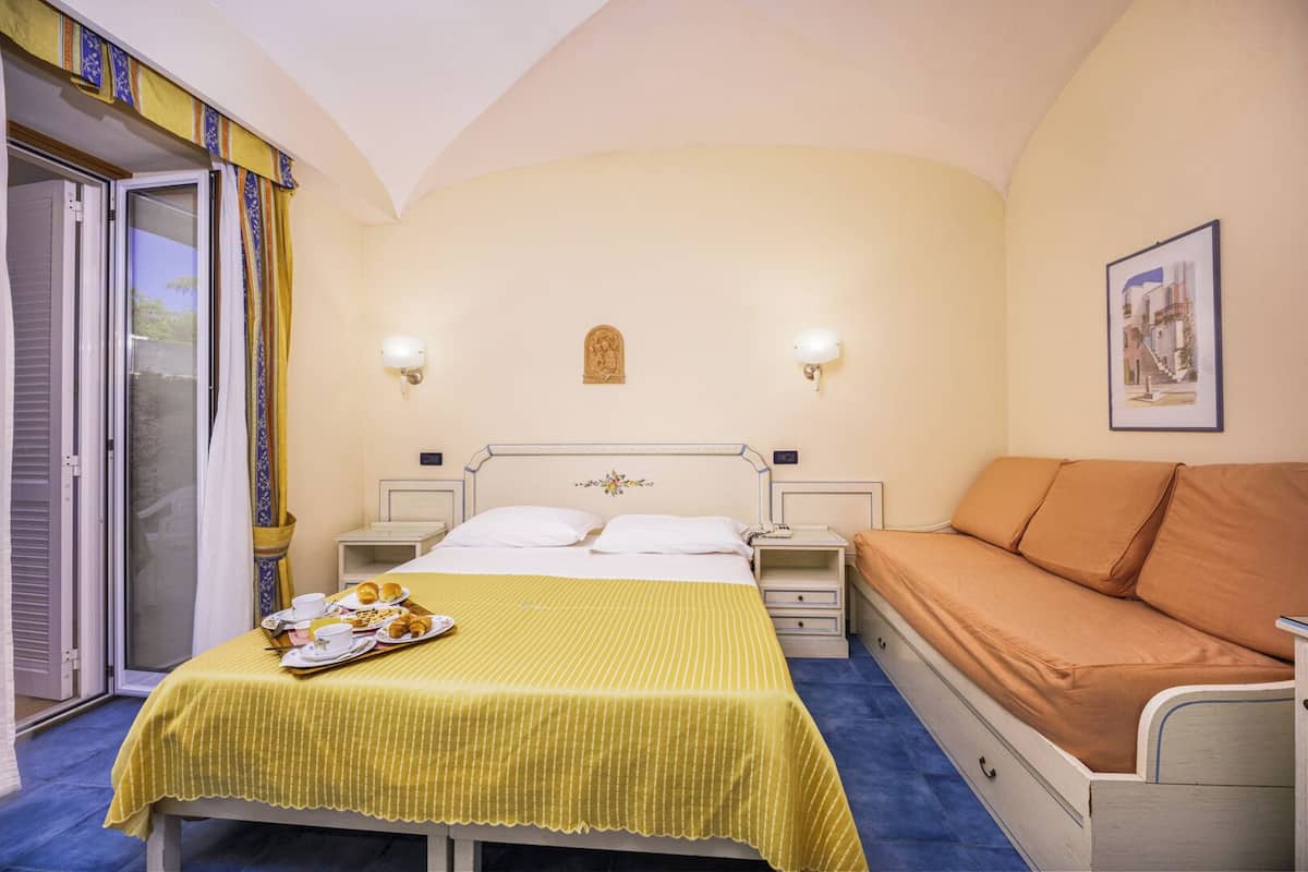Ein Hotelzimmer mit Bett, Nachttischen, einem Sofa, einem Esstisch mit Frühstück und einem Fenster mit Vorhängen.
