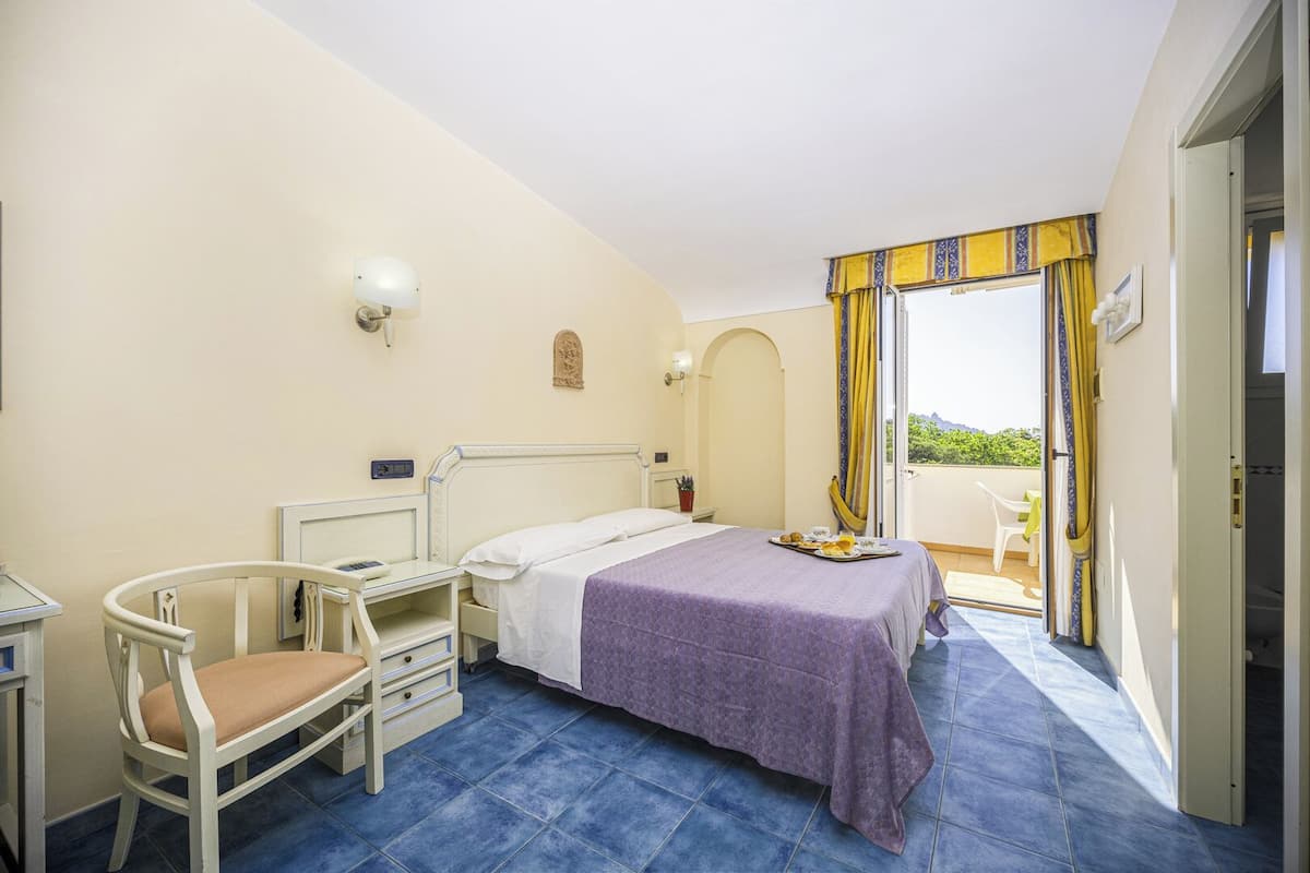Ein Schlafzimmer mit Bett, Nachttisch, Stuhl und einem Balkon mit Vorhängen.