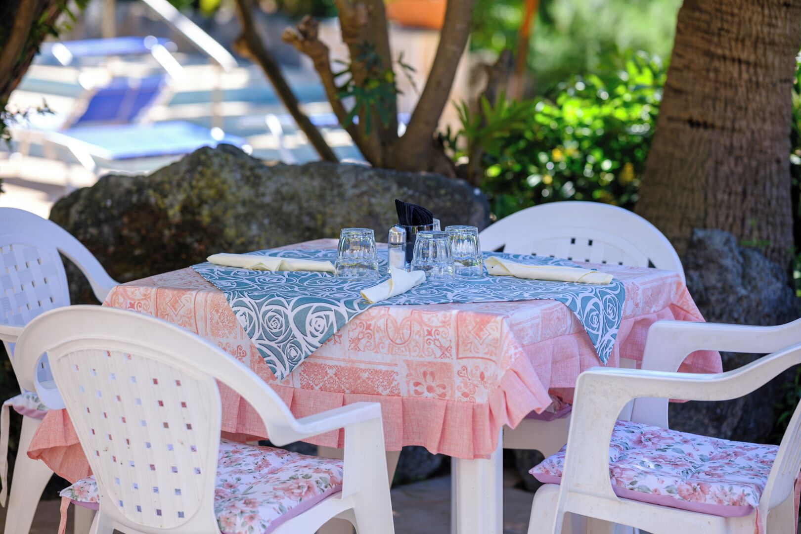 An outdoor dining area with tables covered in patterned tablecloths, white plastic chairs with floral cushions, and a view of a swimming pool in the background.