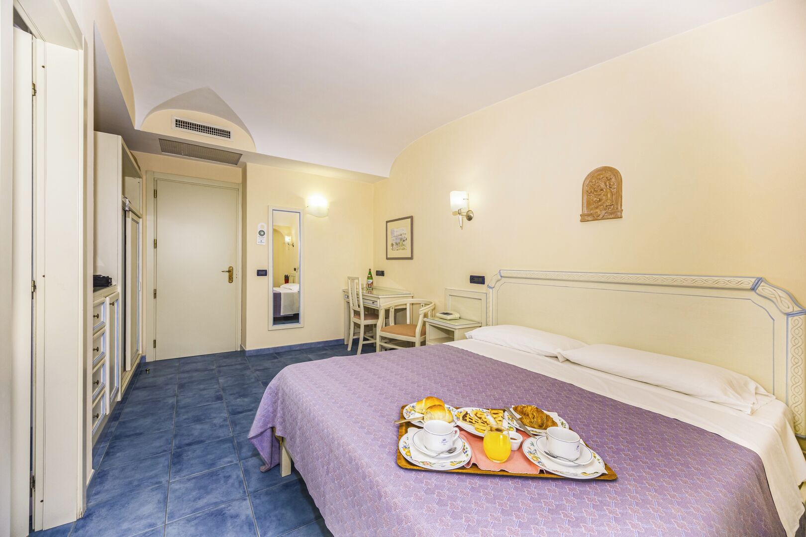 A hotel room with a bed, a desk, a chair, and a tray with breakfast items.