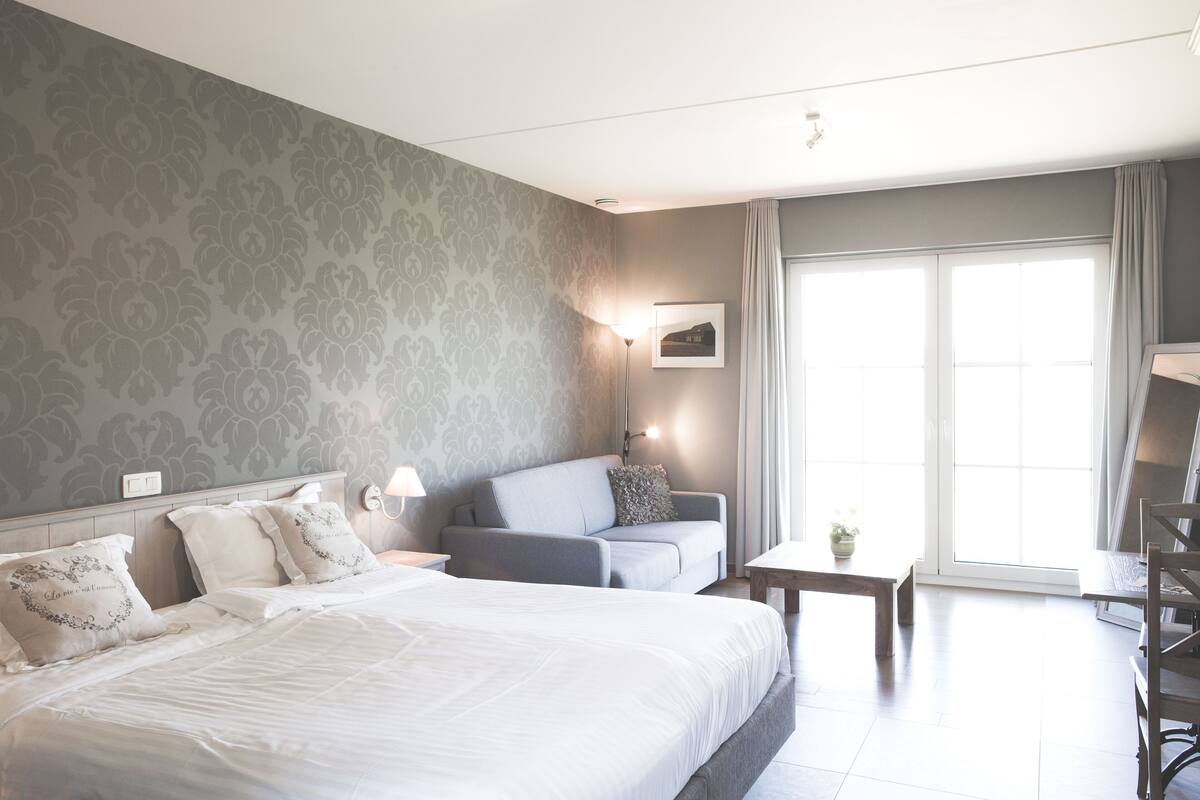 A hotel room with a large bed, a sofa, a small table, and a chair. There is a window with curtains and a lamp on the wall.