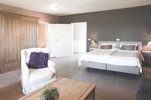 A modern bedroom with a large bed, a white armchair with a purple pillow, a wooden coffee table, and a wardrobe.