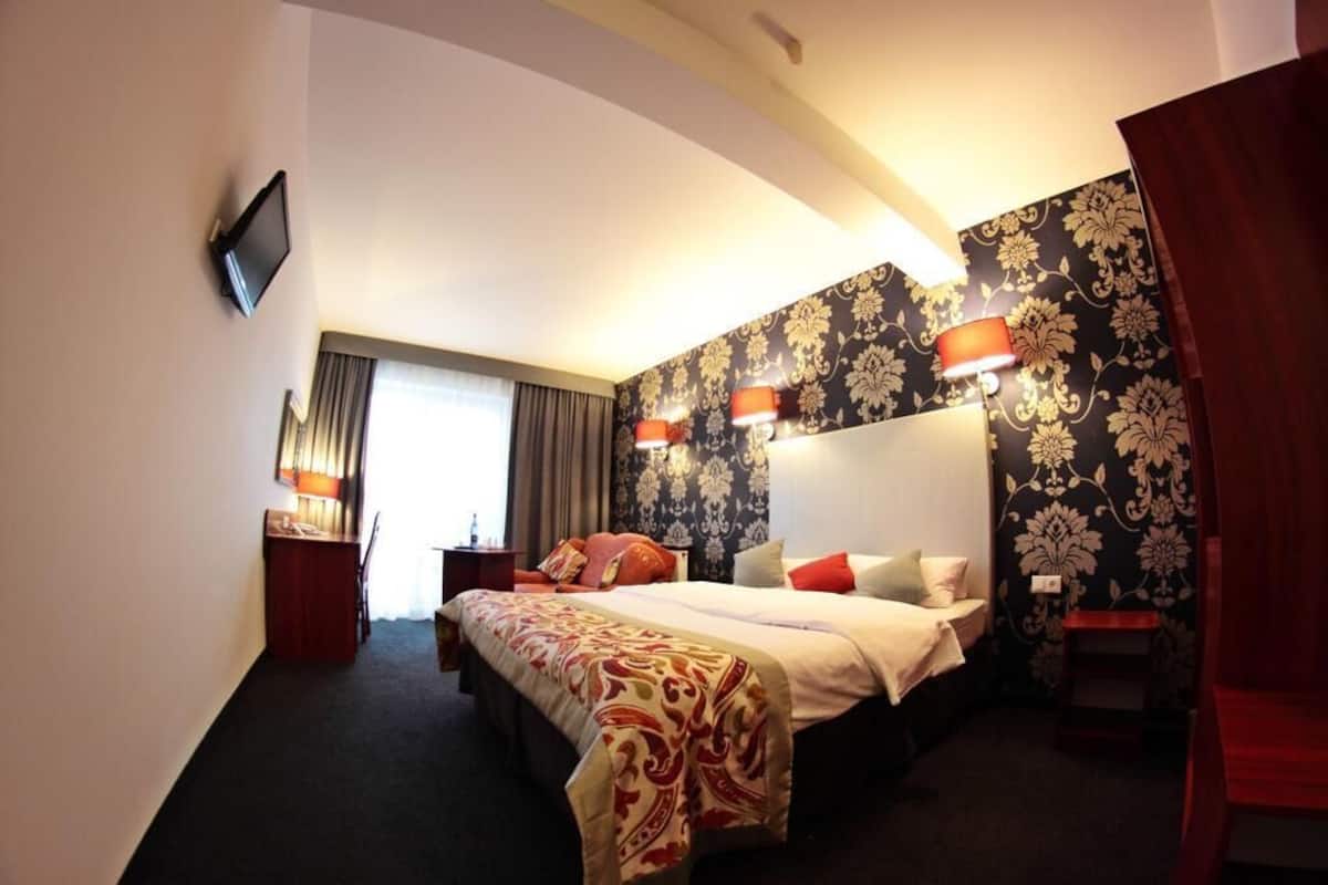 A hotel room with a large bed, two bedside tables, a TV, and a window with curtains.