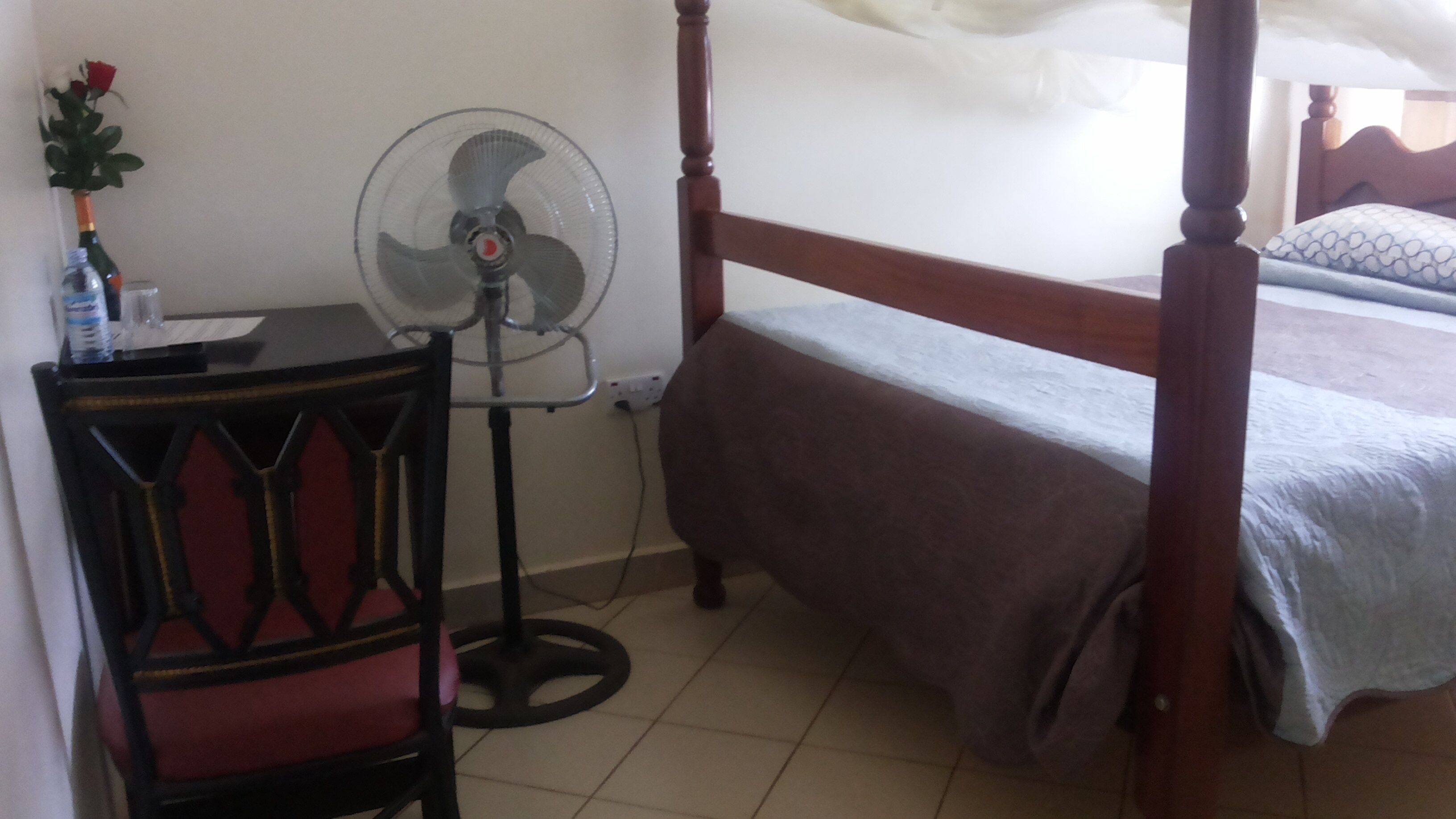 A bedroom with a wooden bed, a bedside table, a fan, and a small table with a bottle and glass.