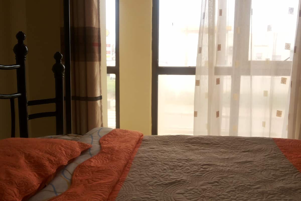 A bedroom with a bed, a window with sheer curtains, and a view of the outside.