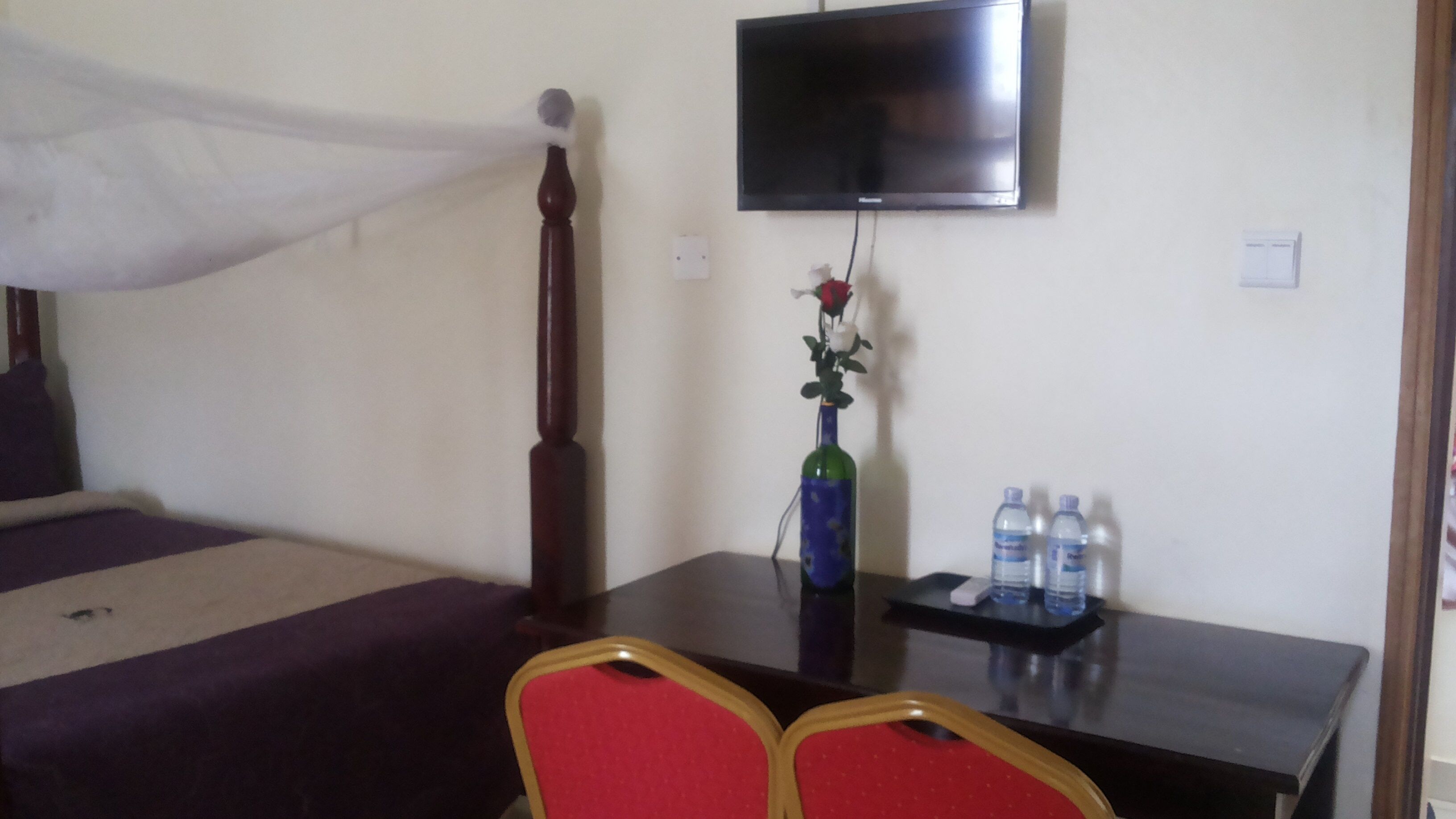 A hotel room with a bed, a desk, a chair, a television, a vase with a flower, and two water bottles.