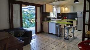 A modern kitchen with a green countertop, a dining area with a sofa and chairs, and a view of tropical foliage through a glass door. - 3-star Lounge des hauts house with ocean & nature view (Etang-Salé les Hauts)