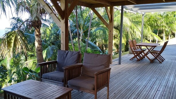 A wooden deck with a covered seating area, including a table and chairs, surrounded by lush tropical vegetation. - 3-star Lounge des hauts house with ocean & nature view (Etang-Salé les Hauts)