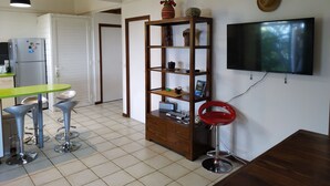 A modern kitchen with a dining area, a TV mounted on the wall, and a wooden shelf with decorative items. - 3-star Lounge des hauts house with ocean & nature view (Etang-Salé les Hauts)