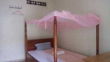 A single bed with a wooden frame and a pink mosquito net. There is a wooden nightstand beside the bed with a white towel on it. A grey pillow is on the bed, and a white towel is on the nightstand.