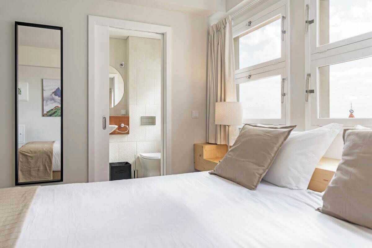 Una camera d'albergo con un letto grande, un comodino con una lampada, uno specchio e vista sulla città dalla finestra.