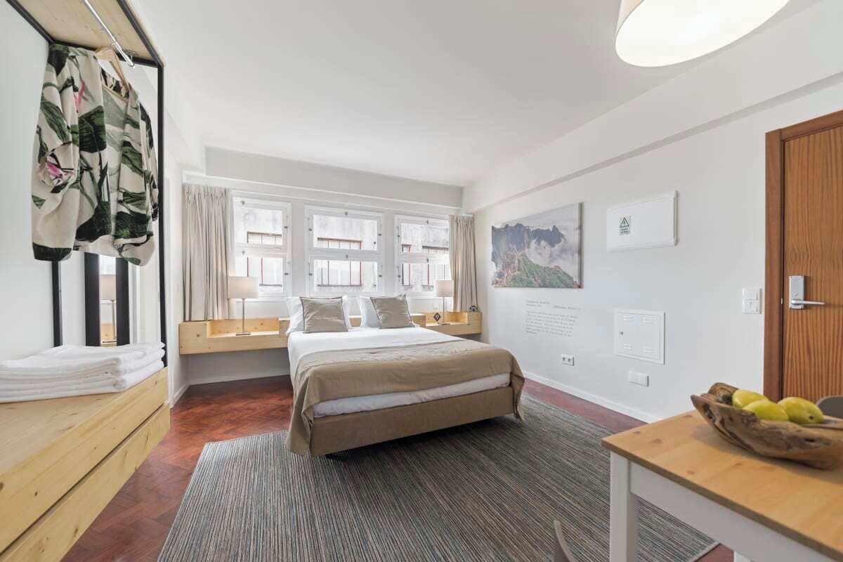 A hotel room with a bed, a desk with a bowl of fruit, a window with curtains, and a wall with a framed picture.