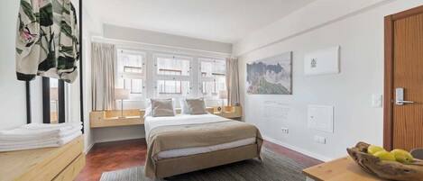 A hotel room with a bed, a desk with a bowl of fruit, a window with curtains, and a wall with a framed picture.