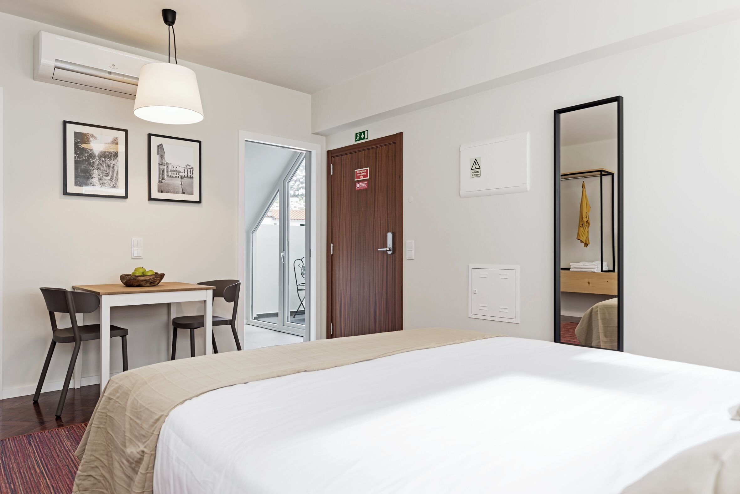 Une chambre d’hôtel avec un lit, un bureau avec deux chaises, une petite table avec des fruits, un miroir et une porte avec un panneau indiquant la sortie.
