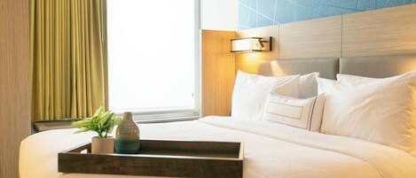 A hotel room with a large bed, a bedside table with a tray, a potted plant, and a window with curtains.