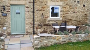 Une petite maison en pierre avec une porte verte, une petite fenêtre et une terrasse aménagée avec du mobilier d’extérieur.